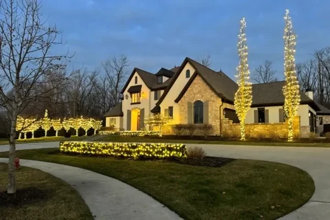 Estate home with warm white roofline lighting, tall spiral lit trees flanking the entry and a lit hedge line along the drive