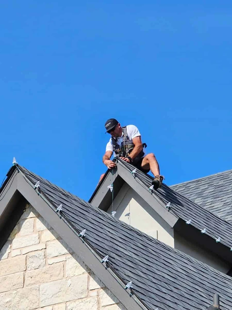 A Sunday installer working on a steep roof ridge, clipping a C9 Christmas light strand into place against a clear blue sky