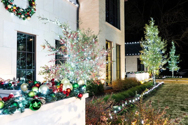 Close detail of a decorated porch with lit garland, oversized ornaments and illuminated greenery beside the front door