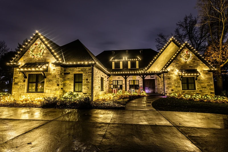 Stone-front home outlined in warm white Christmas lights along every roofline and gable, photographed at night from the driveway