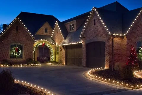 Stone and brick home with warm white roofline lighting, lit wreaths on the gables and a lit walkway to the front door